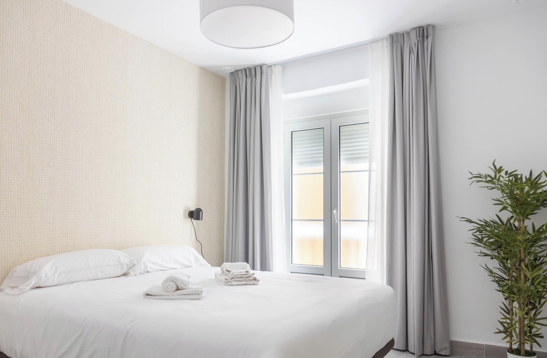 Une chambre d’hôtel avec un lit, une lampe fixée au mur, une fenêtre avec des rideaux et une plante en pot.