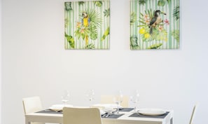 A dining table set with white plates, cutlery, and wine glasses, flanked by chairs. Two tropical-themed paintings adorn the wall above. - Loft Santa Maria by Cadiz Time (Ciudad de Cádiz)