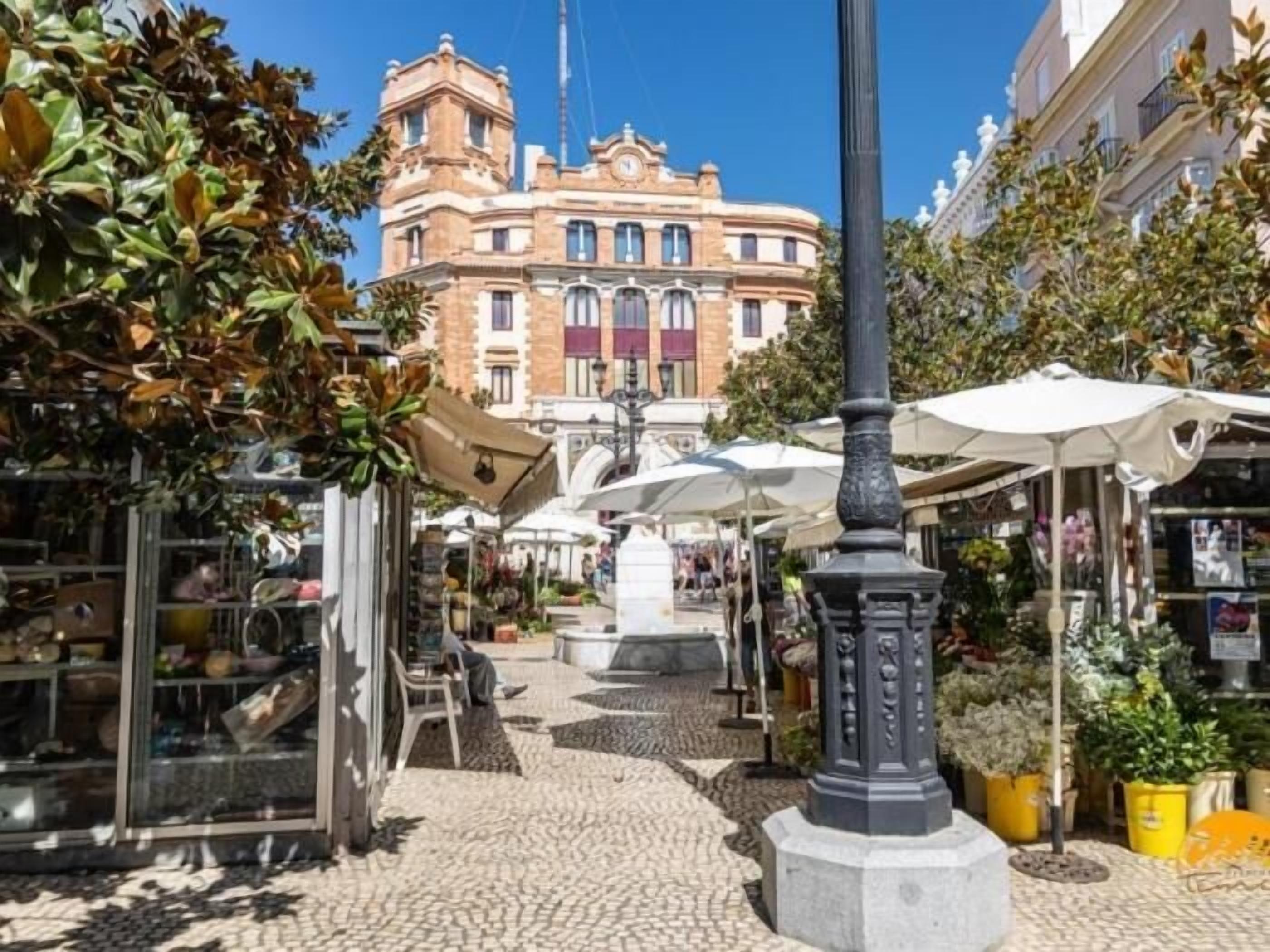 Un carrer empedrat amb botigues, una font al centre i un edifici històric al fons.