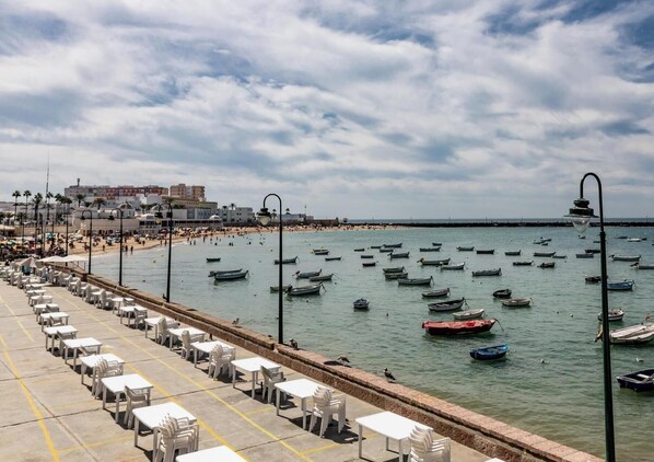 Eine Seepromenade mit Reihen aus weißen Tischen und Stühlen, ein Strand im Hintergrund und zahlreiche Boote auf dem Wasser.