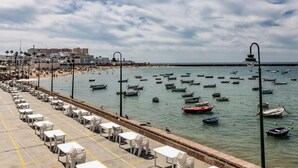 Uma área costeira com um calçadão repleto de mesas e cadeiras, barcos na água e edifícios ao fundo.