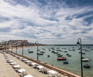 Eine Seepromenade mit Reihen aus weißen Tischen und Stühlen, ein Strand im Hintergrund und zahlreiche Boote auf dem Wasser.