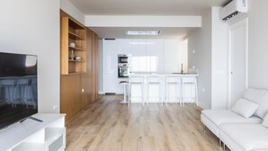 Uma sala de estar moderna com uma TV de tela plana, prateleiras de madeira, um sofá branco e uma cozinha ao fundo.