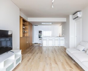 Ein modernes Wohnzimmer mit Flachbildfernseher, Holzregalen, einem weißen Sofa und einer Küche im Hintergrund.