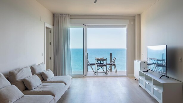 Uma sala de estar moderna com sofá, TV e varanda com vista para o mar.