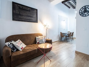 Ein Wohnzimmer mit braunem Sofa, einem runden Couchtisch, einer an der Wand befestigten Periodentafel, einer Uhr und einem Essbereich mit Stühlen und einem Tisch.
