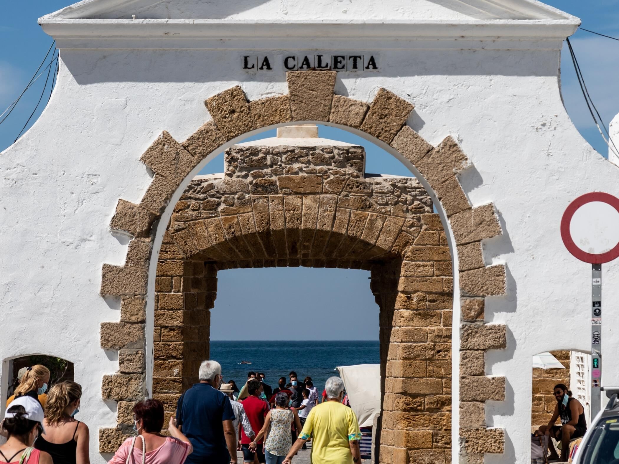 Un arco con la inscripción 'LA CALETTA' en la parte superior, que da paso a una zona costera con gente caminando.