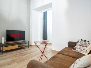 Ein modernes Wohnzimmer mit Flachbildfernseher, einem kleinen Holz-TV-Schrank, einem roten Metall-Beistelltisch, einem braunen Sofa und einem Fenster mit Vorhängen.