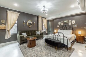 Un dormitorio con cama, sofá, una mesita y un banco. Hay apliques de pared, un espejo y cuadros enmarcados.
