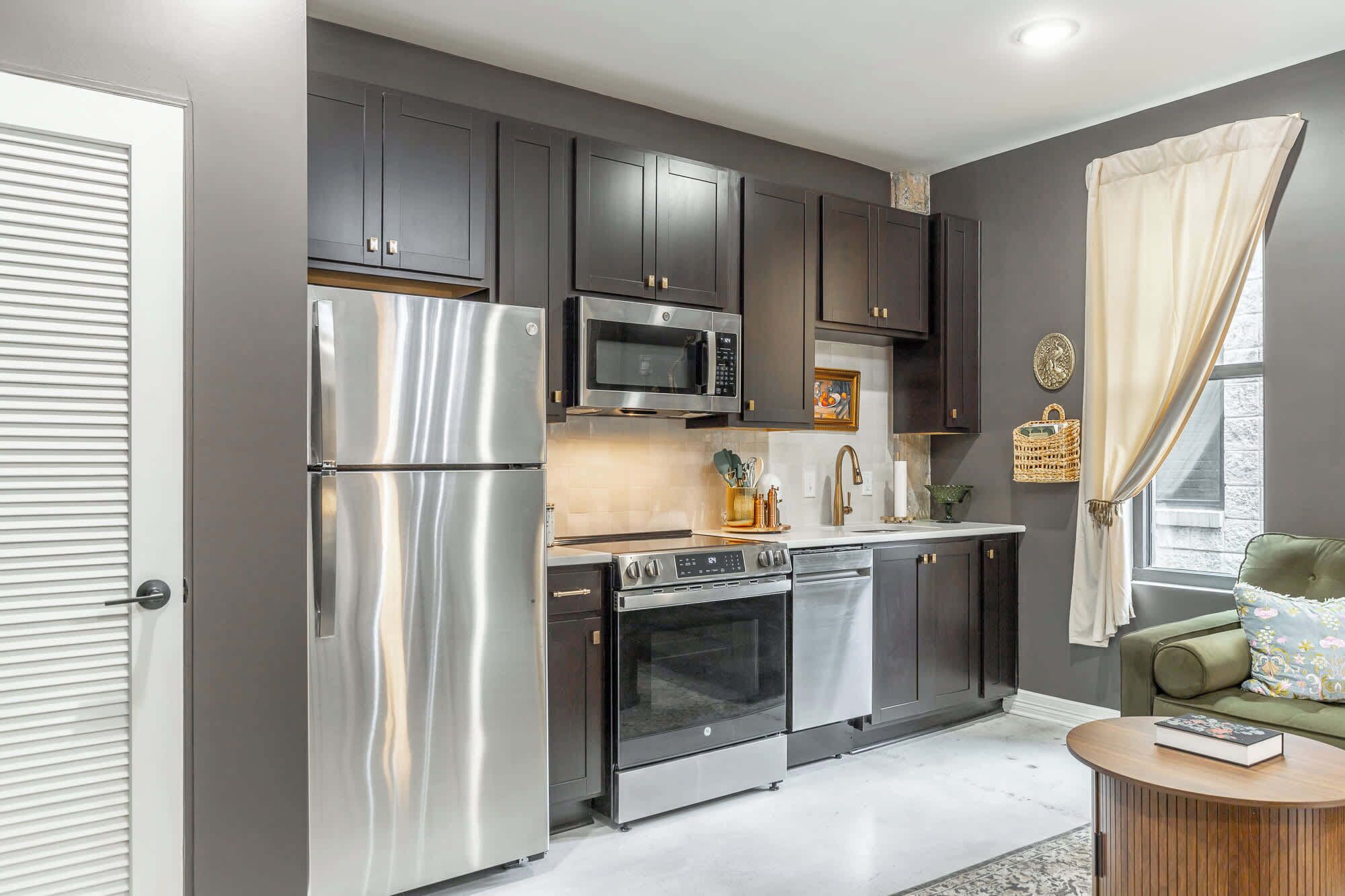A modern kitchen with stainless steel appliances, dark cabinetry, and a window with curtains.