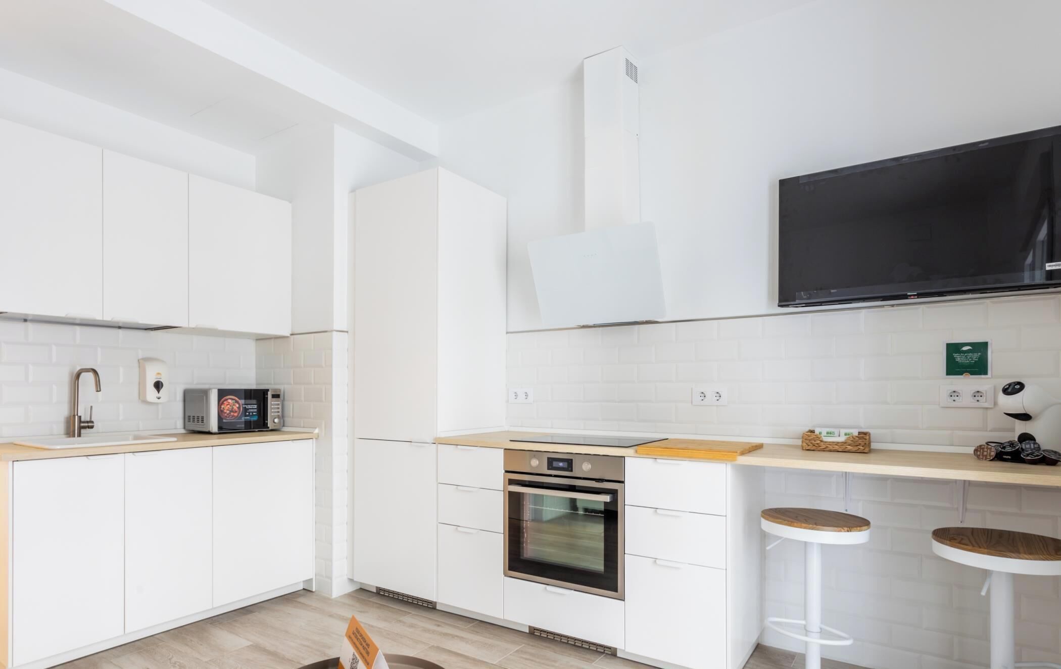 A modern kitchen with white cabinets, a microwave, an oven, and a large flat-screen TV mounted above the counter.