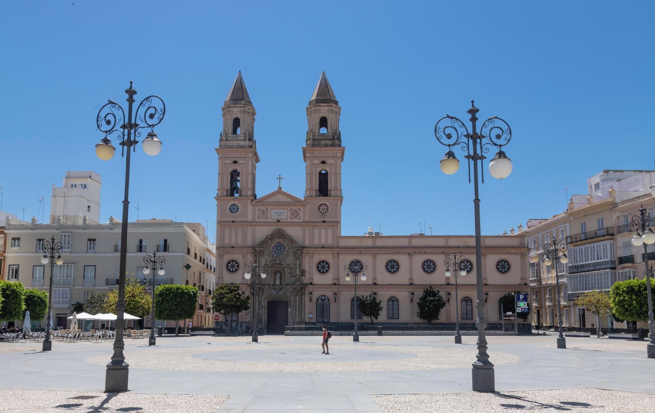 Um edifício histórico com duas torres, uma entrada central e uma praça pavimentada com postes de iluminação.