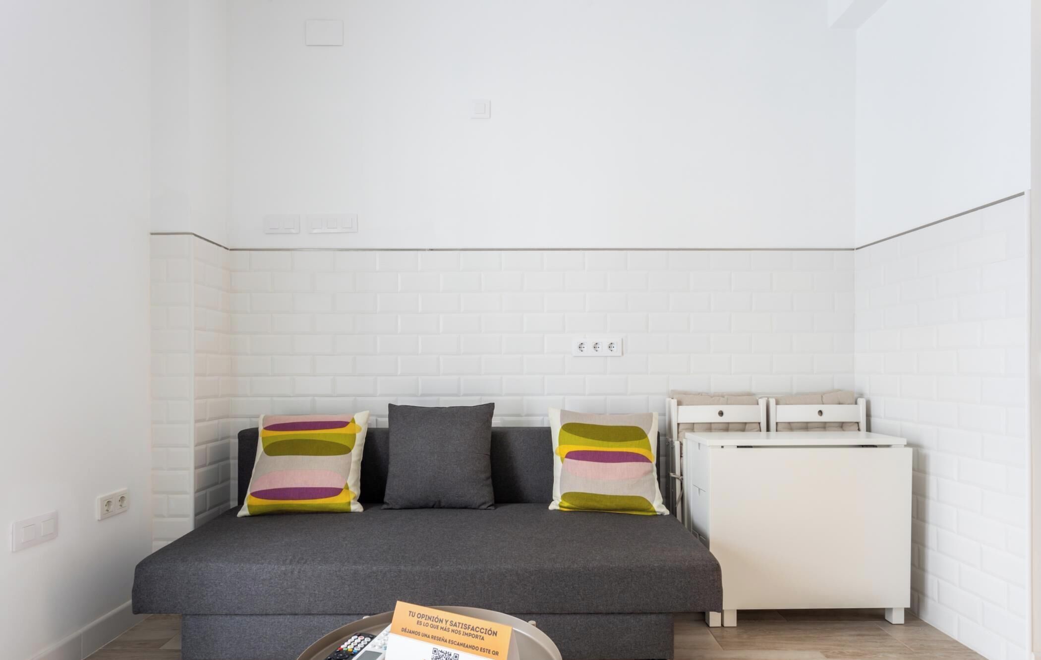 Una habitación pequeña y moderna con un sofá gris, dos almohadas coloridas, una mesita auxiliar blanca y un mueble blanco.