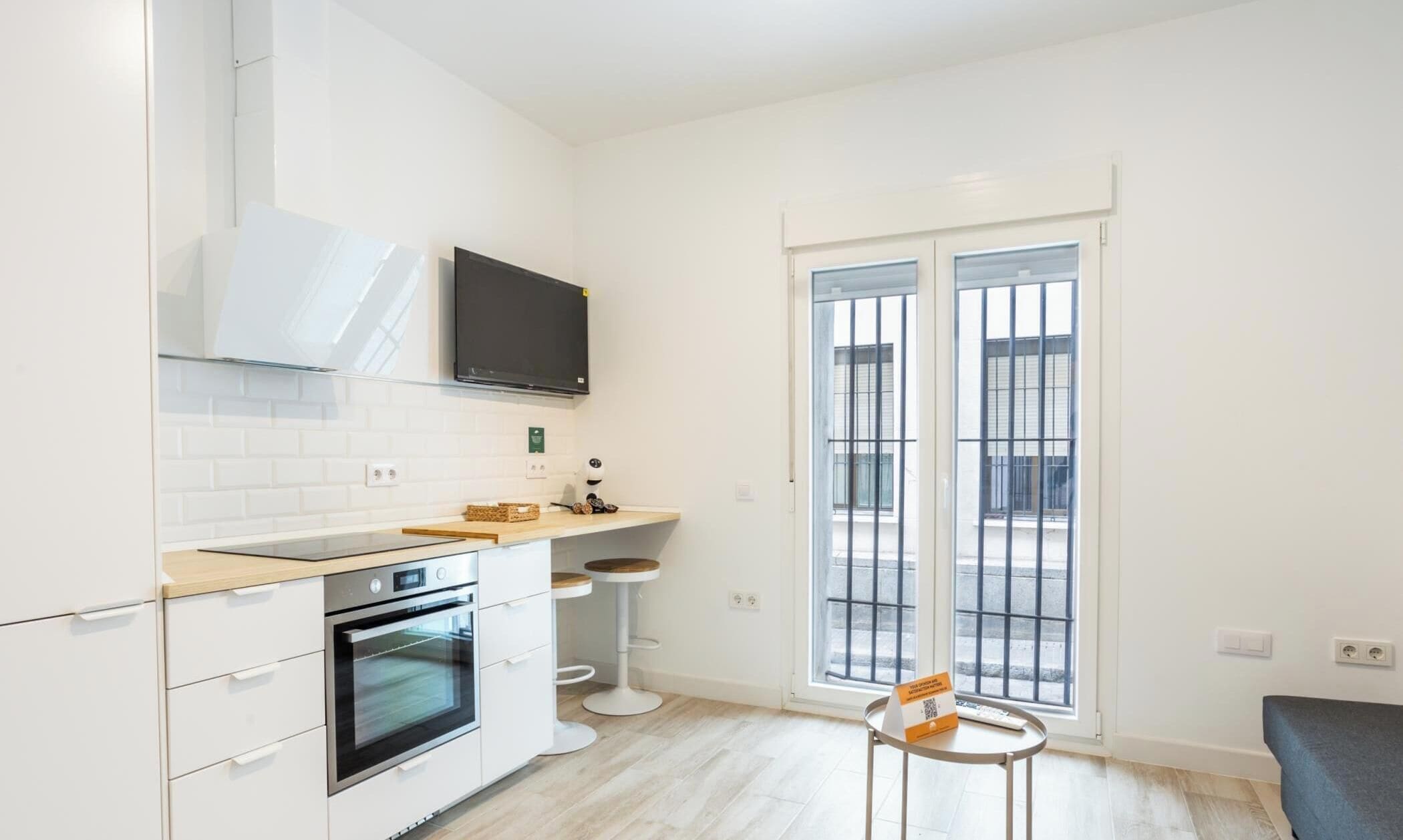 A compact kitchen with a built-in oven, a microwave, a small dining area with stools, and a large window with metal bars.