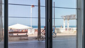 Vista costera a través de una ventana, con playa, una tienda blanca, una estructura metálica y un árbol.