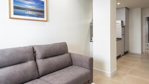Una sala de estar moderna con un sofá gris, un cuadro enmarcado de un paisaje, y una zona de cocina al fondo.
