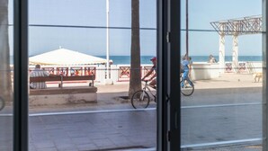Vista costera a través de una ventana, con playa, una tienda blanca, una estructura metálica y un árbol.