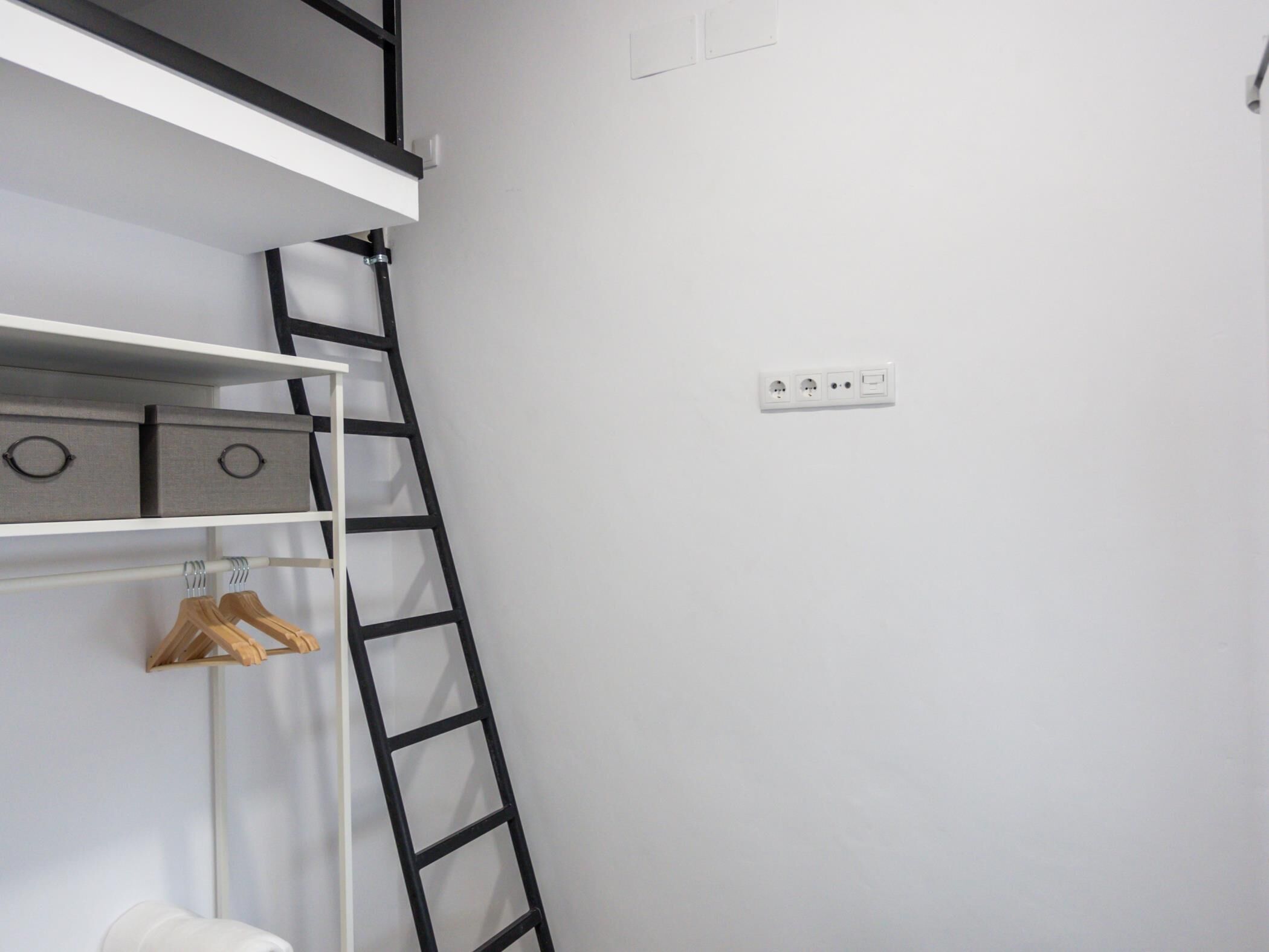 Una habitación blanca con una escalera negra, un estante blanco con cajas grises de almacenamiento y un perchero blanco.
