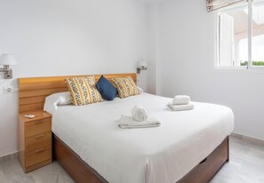 A neatly made bed with white linens, two pillows with floral patterns, a wooden headboard, and a nightstand with a lamp. - Retama Golf by Cádiz Time (Chiclana de la Frontera)