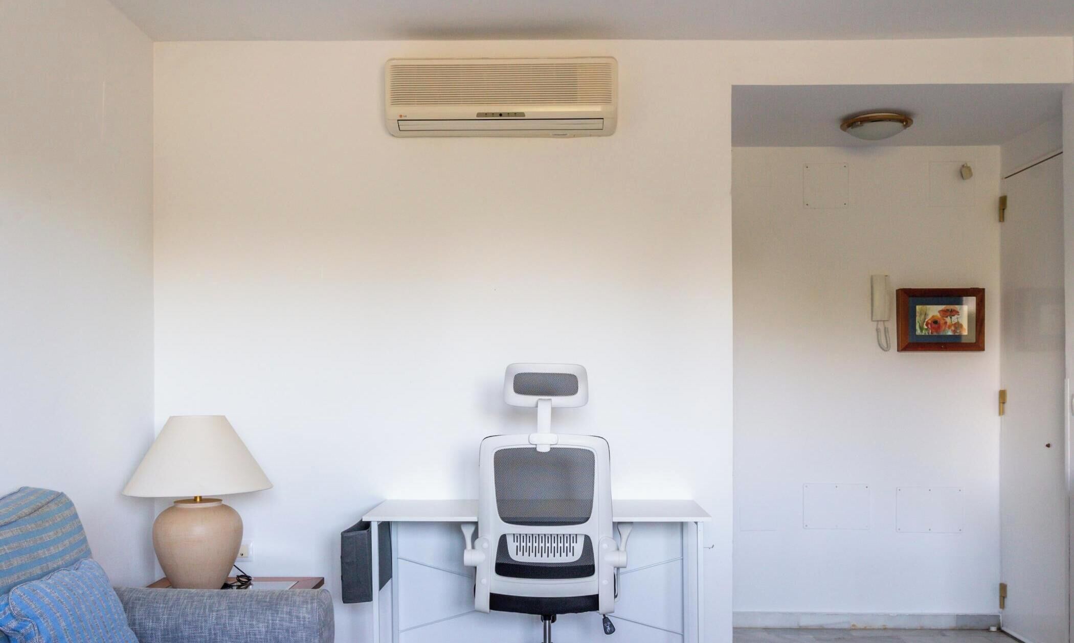 A room with a white desk, a chair, a lamp, and a wall-mounted air conditioner.