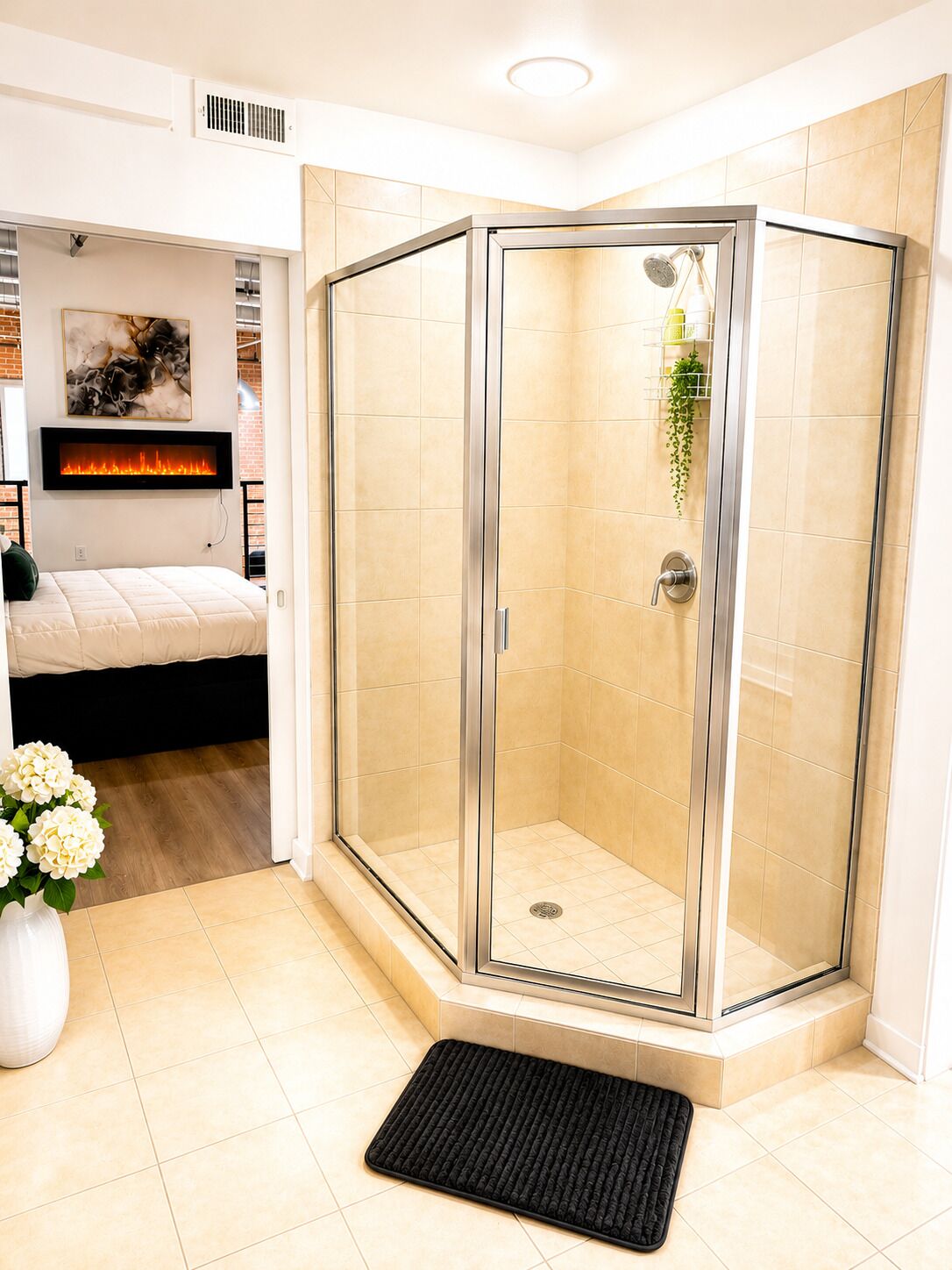A modern bathroom with a glass-enclosed shower, a mounted fireplace, and a bed in the adjacent room.