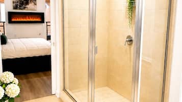 A modern bathroom with a glass-enclosed shower, a mounted fireplace, and a bed in the adjacent room.