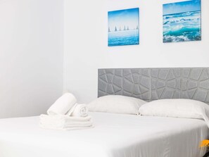 Ein Hotelzimmer mit einem Bett, weißer Bettwäsche, einem Kopfteil mit geometrischem Muster und zwei an der Wand befestigten Bildern vom Meer.