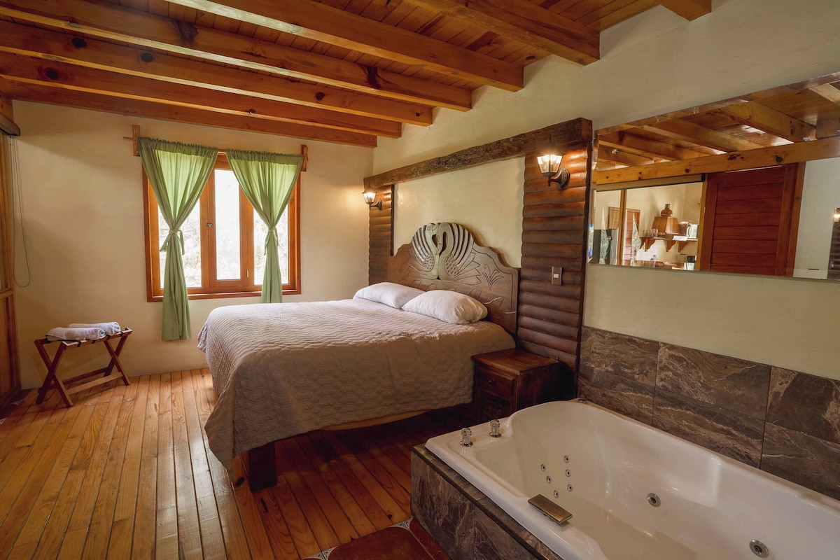 A bedroom with a wooden ceiling, a bed with a headboard, a nightstand, a window with curtains, and a bathroom with a bathtub.