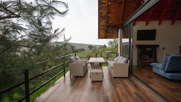 A wooden deck with outdoor furniture, a glass door leading to a room with a fireplace, and a view of trees and hills.