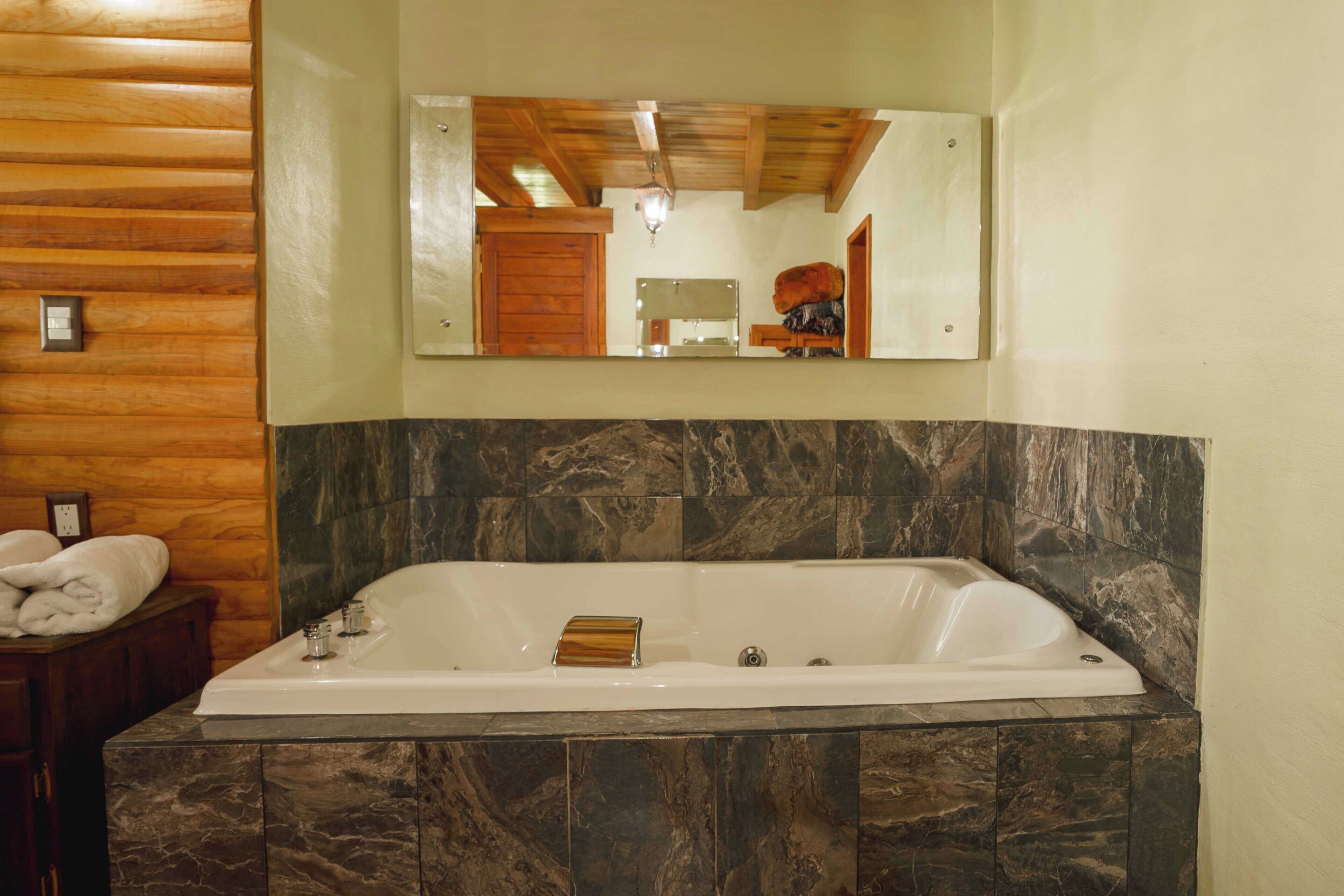 A bathroom with a large bathtub, marble tiles, wooden paneling, and a mirror above the tub.