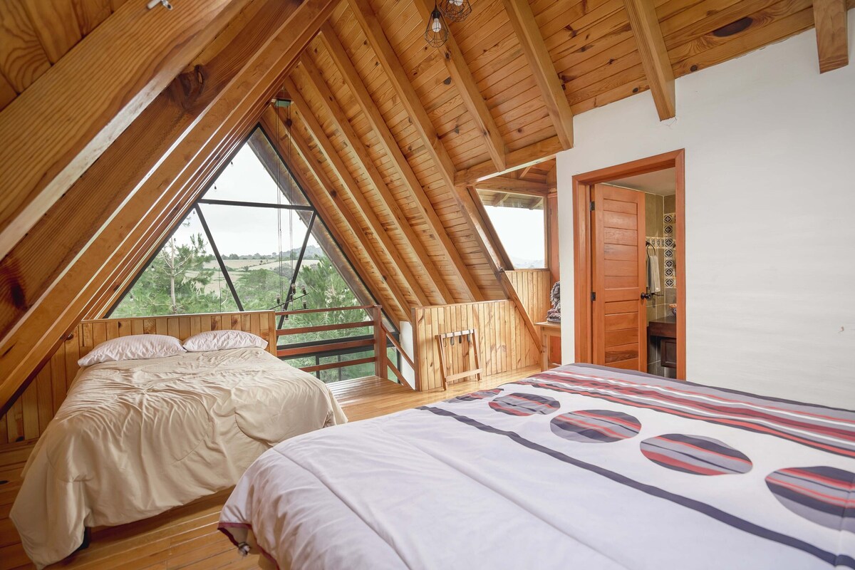 A bedroom with a sloped ceiling, wooden beams, two beds, a window with a view of greenery, and a door leading to a bathroom.