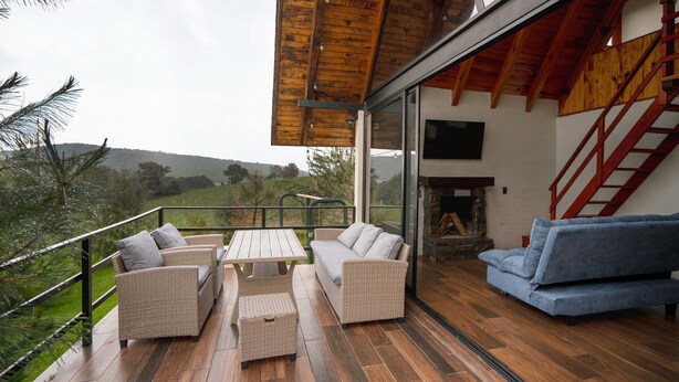 A wooden deck with outdoor furniture, a stone fireplace, and a view of the countryside. - Paseo del Llano Cabañas & Suites (Mazamitla)