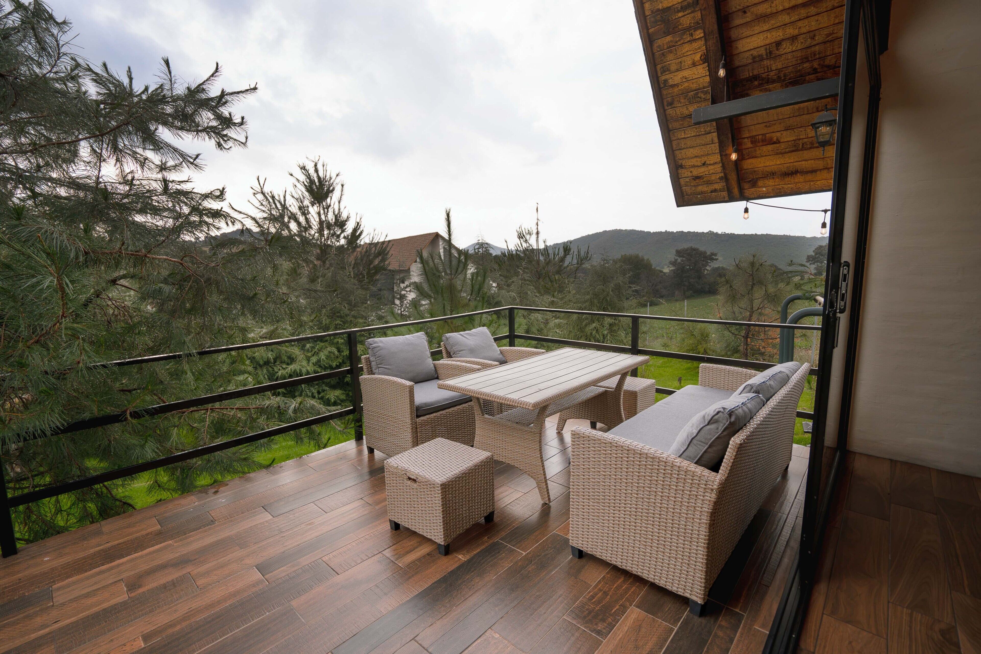 Un balcón con mobiliario de mimbre, piso de madera y vistas a árboles y colinas.