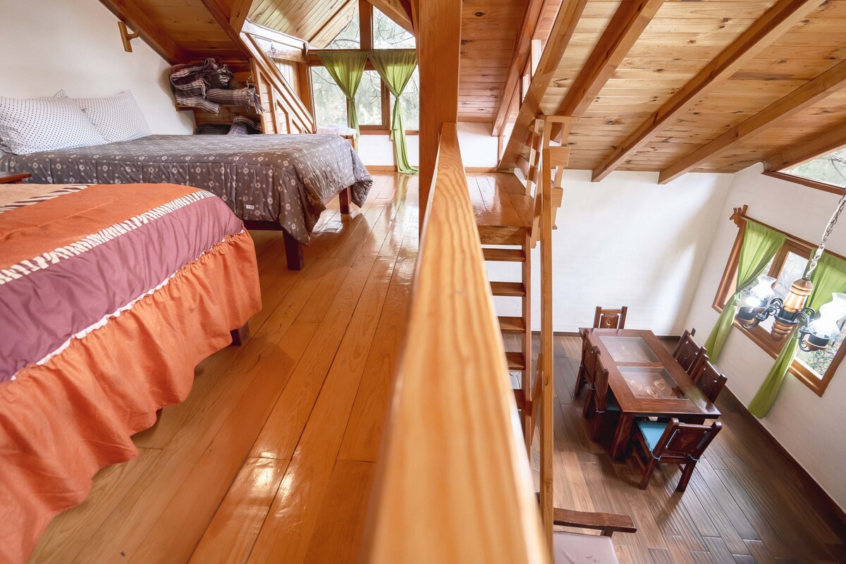A room with a wooden floor, a bed with a patterned bedspread, a dining table with chairs, and a window with curtains.