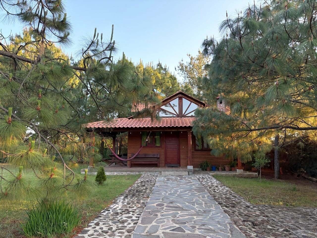 A wooden cabin with a red-tiled roof, surrounded by pine trees and a stone pathway leading to the entrance.