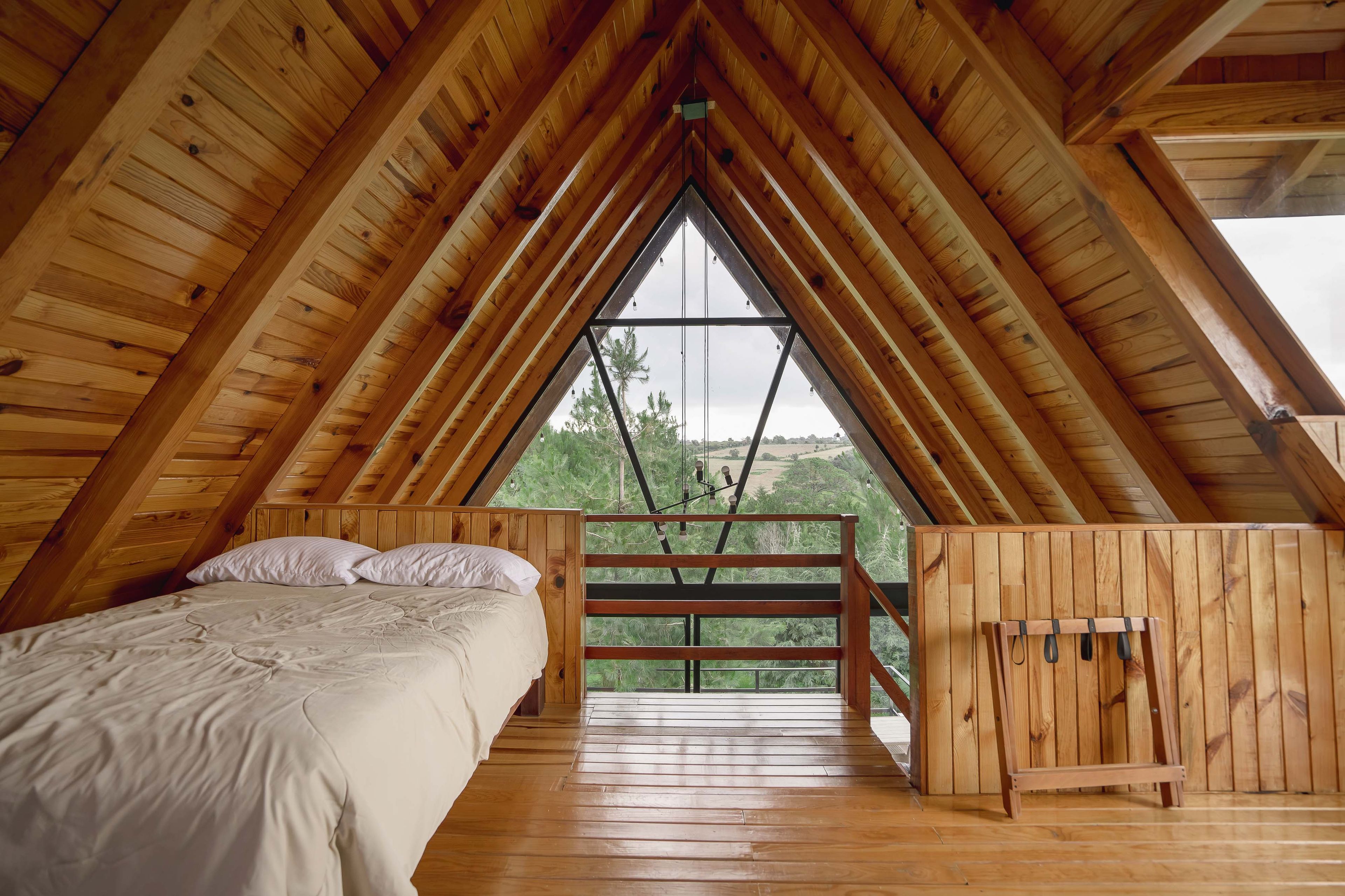 A wooden attic bedroom with a bed, a bench, and a large window offering a view of greenery.