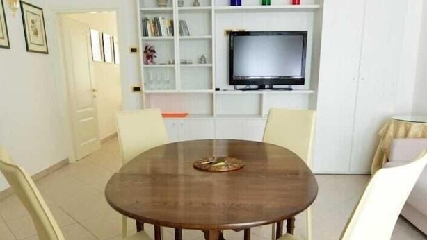 Un comedor con una mesa redonda de madera y cuatro sillas, un televisor en un estante y un armario blanco con estanterías.