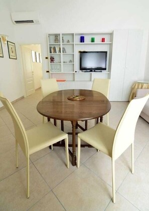Un coin repas comprenant une table ronde en bois et quatre chaises, un téléviseur posé sur une étagère, et un meuble blanc avec des étagères.