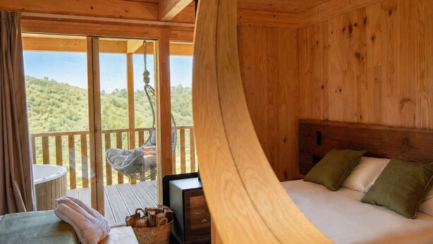 A wooden cabin room with a bed, a small table, a chair, and a view of the outdoors.