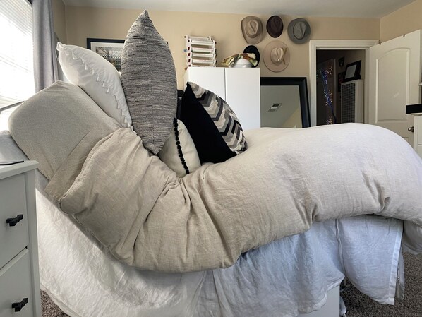 Ein Bett mit vielen Kissen, ein Nachttisch, eine Kommode und eine Wand mit Hüten.