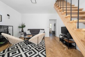 Ein modernes Wohnzimmer mit einer Holztreppe, einem Sofa, zwei Sesseln, einem Fernseher und einem Couchtisch.