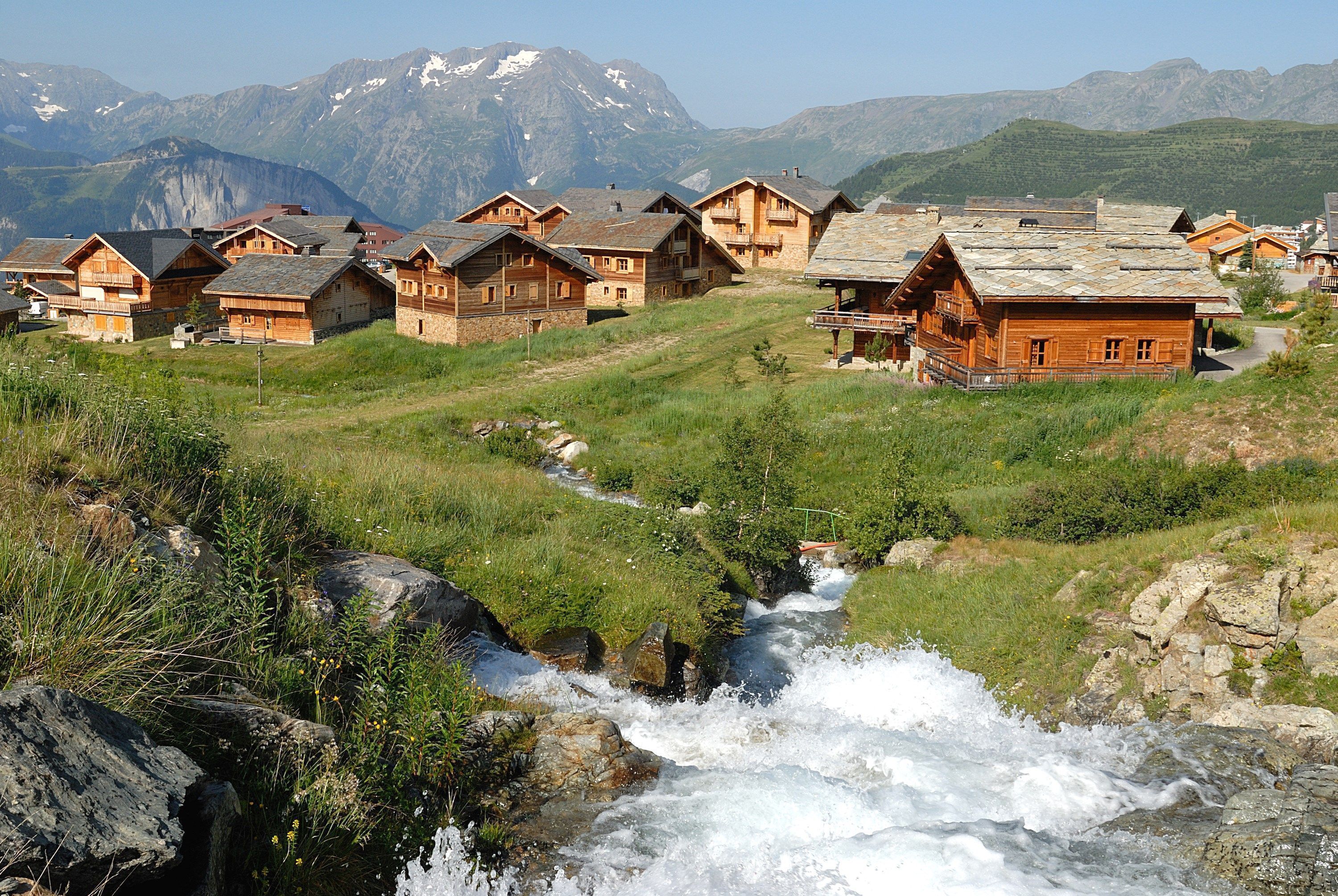 A cluster of wooden houses with a stream flowing through a grassy area, surrounded by mountains.