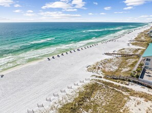 An aerial view of a beach with a long stretch of umbrellas and beach chairs, a wooden boardwalk, and a building with a blue roof. - Sunfish Royale | Private Beach, Billiards, Spa, Pickleball (Destin)