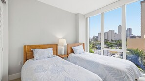 Ein Hotelzimmer mit zwei Betten, einem Nachttisch, einer Lampe und einem großen Fenster mit Blick auf die Stadt.