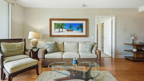 Ein Wohnzimmer mit einem Rattan-Sofa, einem Glastisch, einem Rattan-Sessel und einem gerahmten Strandbild an der Wand.