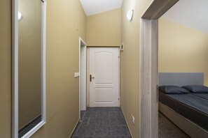 Ein schmaler Flur mit einer Tür, einem Bett mit Kopfteil und einer Wandleuchte.