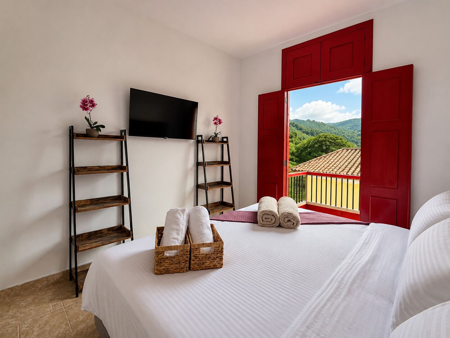 A hotel room with a bed, a TV, a balcony with red shutters, and a view of greenery.