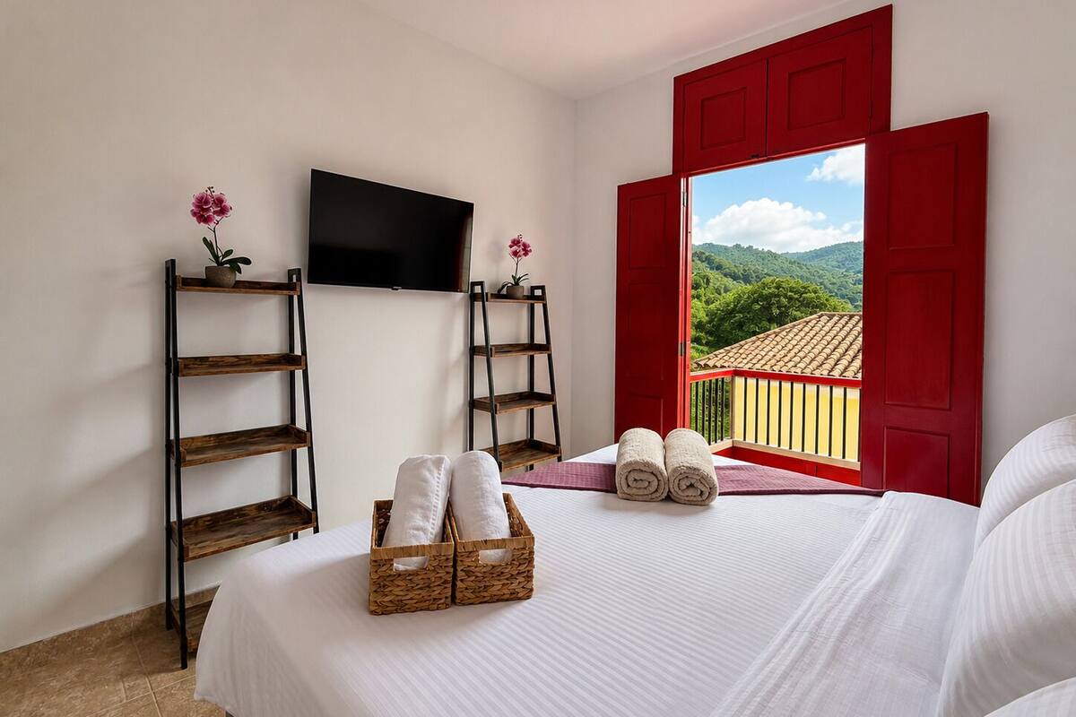 A hotel room with a bed, a TV, a balcony with red shutters, and a view of greenery.
