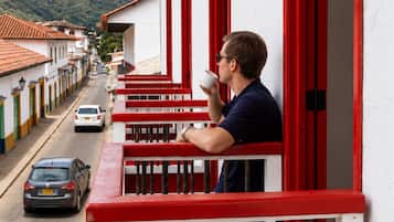 Un hombre en un balcón con barandillas rojas, mirando una calle con coches aparcados y edificios con puertas rojas.
