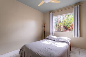 Um quarto com cama, ventoinha no teto, janela com cortinas e vista para árvores lá fora.